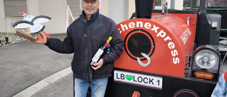 Ein älterer Mann steht neben einem roten Zug, hält eine Flasche Wein und einen Vogel. Der Zug hat ein Schild mit der Aufschrift 'chenexpress'.