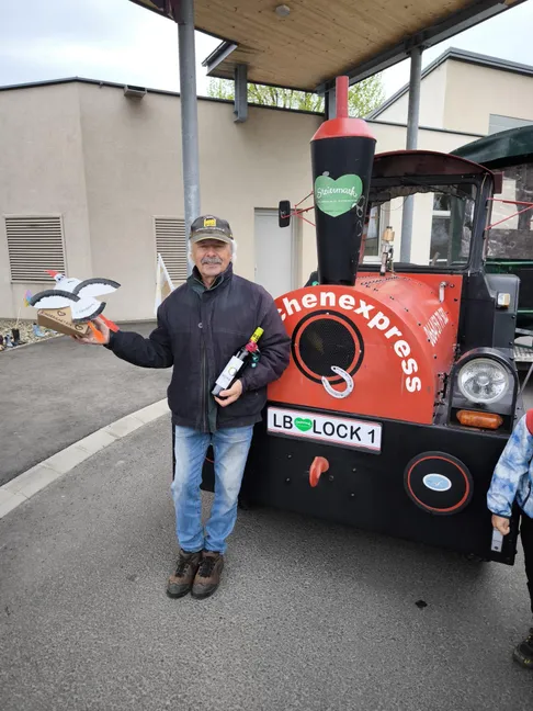 Ein Mann hält eine Weinflasche vor einem roten Zug, mit einem Kind in der Nähe. Der Zug trägt den Aufdruck 'chenexpress'.