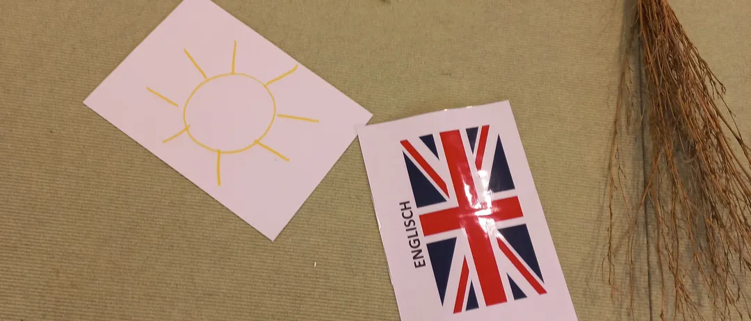 Ein gelbes Papiersonne mit Strahlen, eine weiße Papiersonne, eine Union Jack Flagge und ein Besen liegen auf einer beigen Oberfläche.
