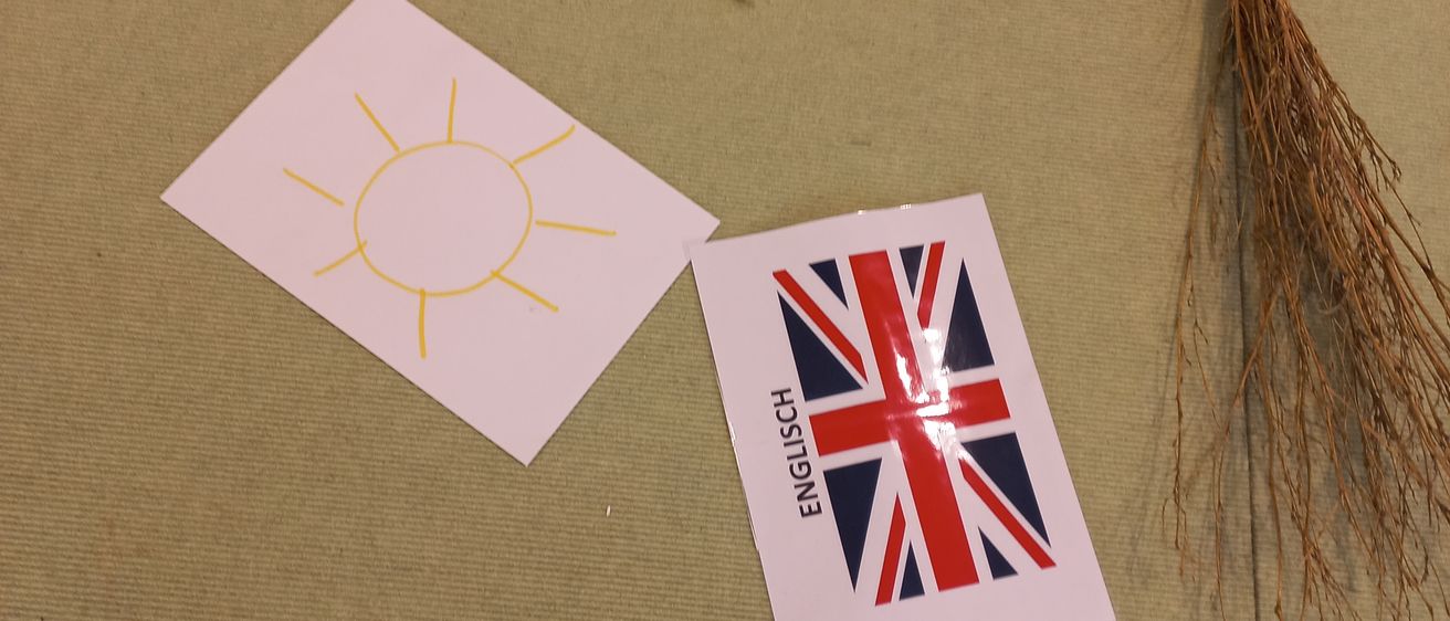 Ein gelbes Papiersonne mit Strahlen, eine weiße Papiersonne, eine Union Jack Flagge und ein Besen liegen auf einer beigen Oberfläche.