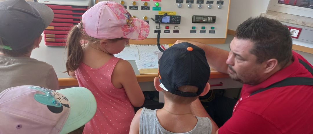 Ein Mann in einem roten Shirt zeigt Kindern, wie man ein Bedienfeld in einem Raum mit Fenster und Drucker im Hintergrund benutzt.