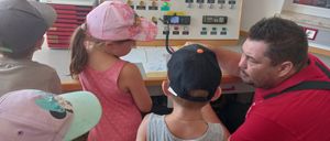 A man in a red shirt is teaching children how to use a control panel in a room with a window and a printer in the background.