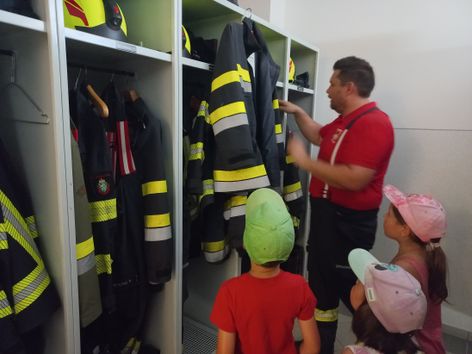 Ein Mann in einem roten Shirt zeigt Kindern Feuerwehruniformen in einem Umkleideraum. Die Uniformen hängen an Kleiderbügeln, und die Kinder tragen Mützen.