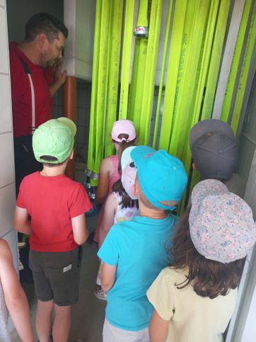 Eine Gruppe von Kindern mit Mützen steht in einem Raum mit grünen Bannern, und ein Mann in einem roten Hemd steht hinter ihnen.