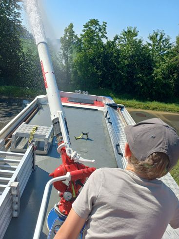 Ein Feuerwehrmann bedient eine Feuerwehrspritze auf einem Boot und sprüht Wasser in die Luft. Das Boot ist am Straßenrand geparkt, mit Bäumen und Büschen im Hintergrund.