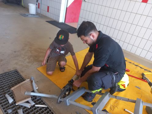Ein Mann in Uniform bringt einem kleinen Jungen in einer Garage bei, wie man ein Werkzeug auf einem gelben Teppich benutzt.