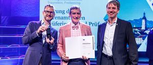 Drei Männer stehen auf einer Bühne. Der Mann in der Mitte hält ein Zertifikat und lächelt. Der Mann auf der linken Seite hält eine Medaille. Sie tragen alle formelle Kleidung. Hinter ihnen befindet sich eine große Leinwand mit Text.