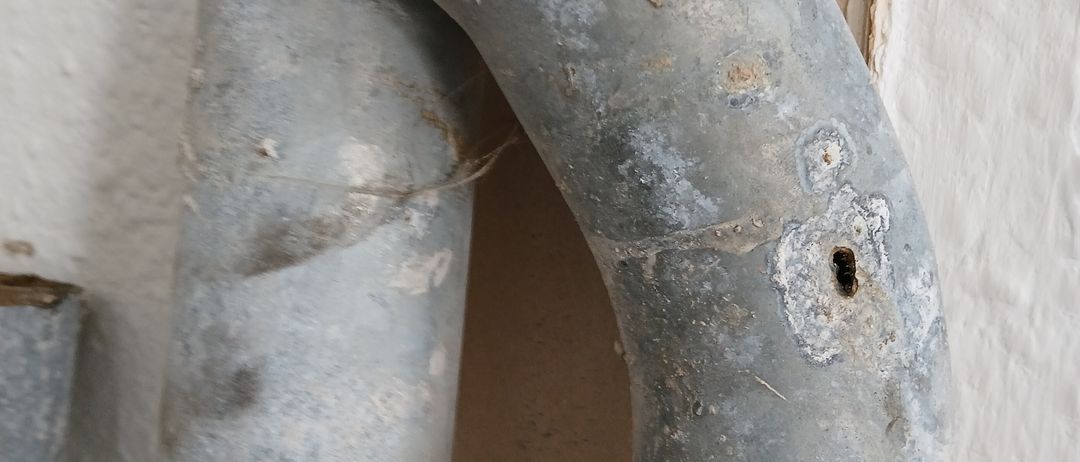 Nahaufnahme eines rostigen Metallrohrs mit einem Loch und Korrosionsspuren. Das Rohr ist an einer weißen Wand befestigt.