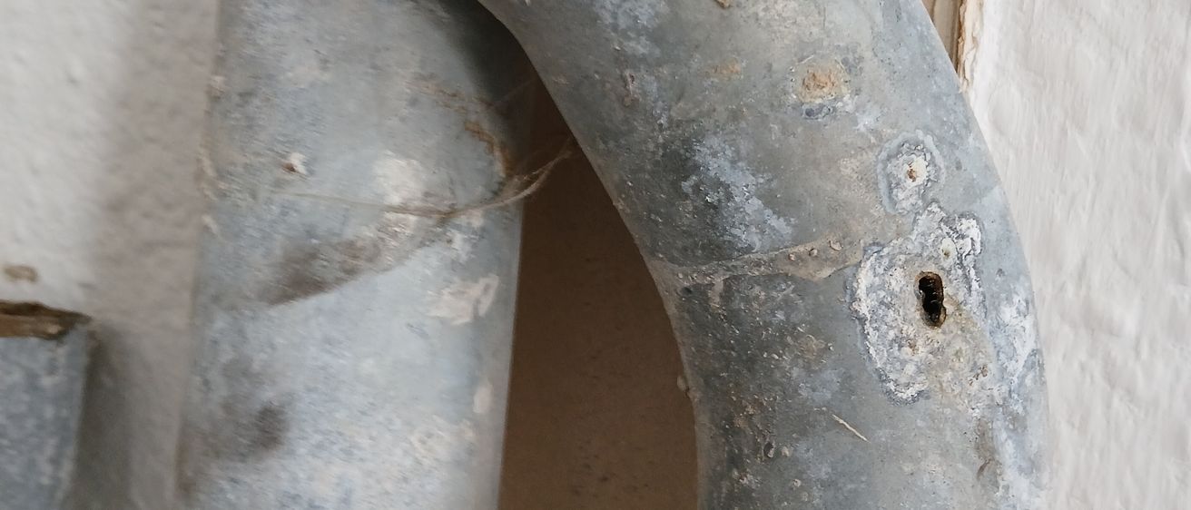 Nahaufnahme eines rostigen Metallrohrs mit einem Loch und Korrosionsspuren. Das Rohr ist an einer weißen Wand befestigt.