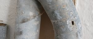 Nahaufnahme eines rostigen Metallrohrs mit einem Loch und Korrosionsspuren. Das Rohr ist an einer weißen Wand befestigt.