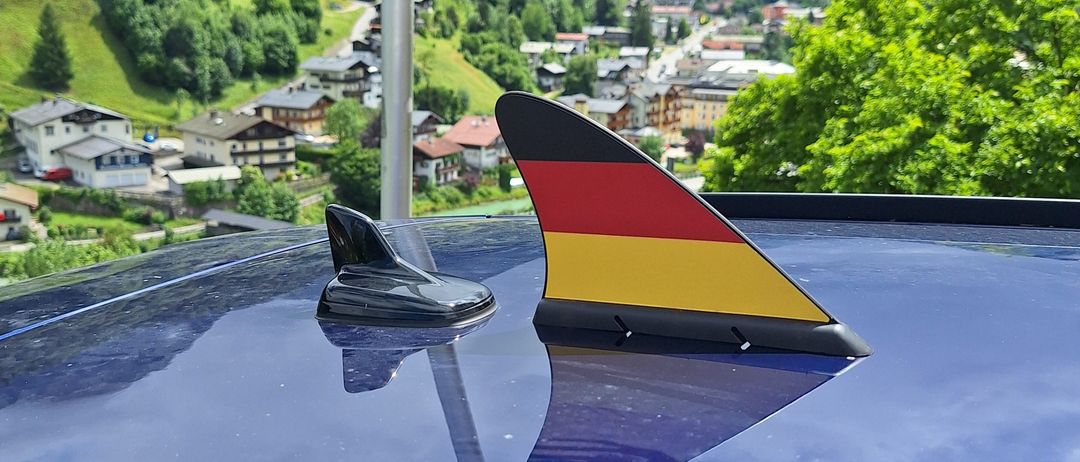 A German flag antenna on a car is reflected in the blue surface, with mountains and houses in the background.