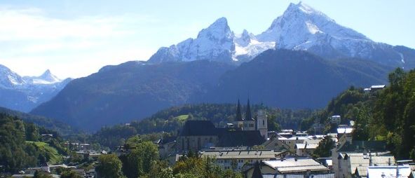 Ein Dorf mit vielen Gebäuden, eingebettet zwischen Bergen und umgeben von Bäumen. In der Ferne befindet sich eine Kirche und eine Bergkette mit Schnee.