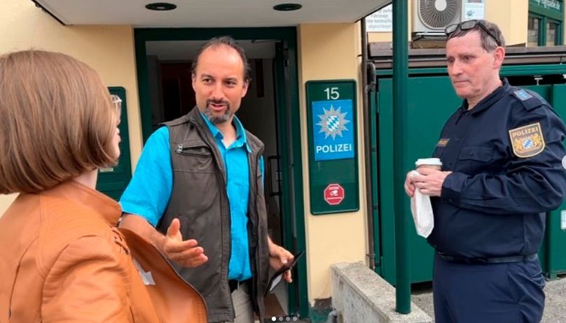 Ein Mann in einem blauen Shirt spricht mit einer Frau, während ein Polizeibeamter zusieht. Der Polizeibeamte hält einen Kaffee und eine Serviette. Ein blaues Schild mit der Nummer 15 und einem Logo ist sichtbar.