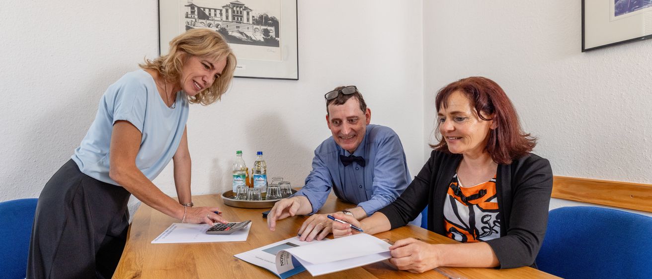 Drei Personen sitzen an einem Holztisch. Zwei Frauen und ein Mann. Der Mann trägt einen Fliege. Ein Taschenrechner, ein Stift und Papiere sind auf dem Tisch. Ein Tablett mit Flaschen und Gläsern steht auf dem Tisch. Ein gerahmtes Bild ist an der Wand.