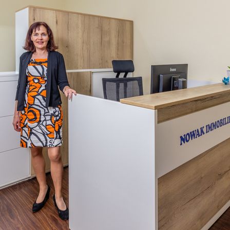Eine Frau in einem gemusterten Kleid und schwarzen Schuhen steht lächelnd hinter einem NOWAK IMMOBILIEN Empfangstresen. Hinter ihr befindet sich ein Monitor und ein Stuhl.