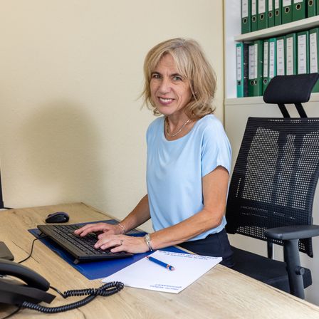 Eine Frau sitzt an einem Schreibtisch, lächelt und tippt auf einem Computer. Sie hat ein Telefon und Papiere vor sich.