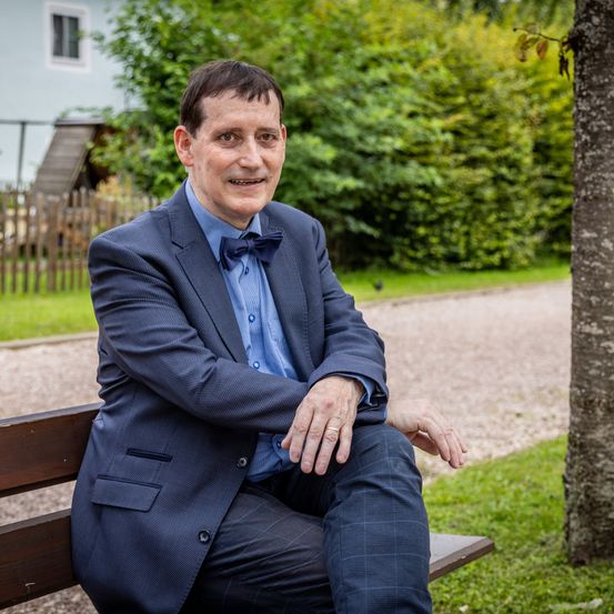 Ein Mann in Anzug und Fliege sitzt lächelnd auf einer Bank und schaut in die Kamera. Dahinter ein Rasen und ein Baum. Im Hintergrund ein Haus und ein Holzzaun.