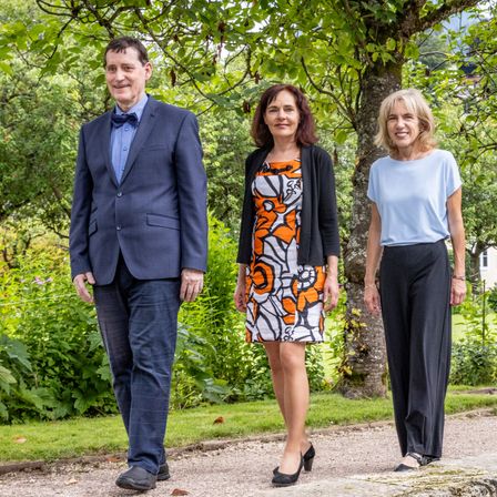 Drei Menschen stehen lächelnd in einem Garten für die Kamera. Ein Mann in Anzug und eine Frau in einem Kleid stehen links, mit einer Frau in einem blauen Oberteil rechts.
