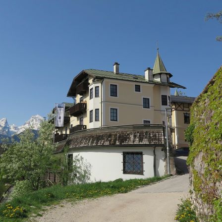 Ein großes Gebäude mit grünem Dach und weißen Wänden steht neben einer unbefestigten Straße, umgeben von Grün und Bergen in der Ferne.