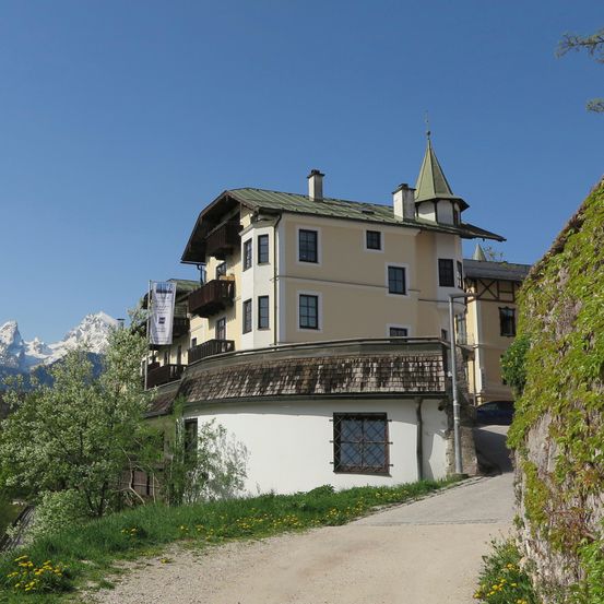 Ein großes Gebäude mit grünem Dach und weißen Wänden steht neben einer unbefestigten Straße, umgeben von Grün und Bergen in der Ferne.