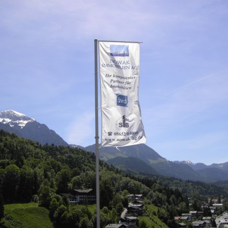 Ein Banner von NOWAK Immobilien AG steht vor einer Kulisse aus Bergen und einem Dorf. Das Banner zeigt das Logo und die Kontaktdaten des Unternehmens.