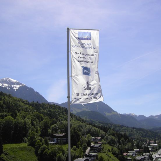 Ein Banner von NOWAK Immobilien AG steht vor einer Kulisse aus Bergen und einem Dorf. Das Banner zeigt das Logo und die Kontaktdaten des Unternehmens.