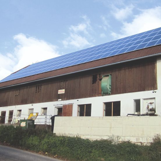 Ein Gebäude mit Solarpanelen auf dem Dach. Es ist überwiegend braun und weiß mit einigen Fenstern. Ein grüner Müllcontainer steht davor.