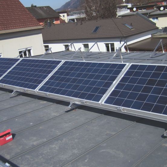 Ein Dach mit mehreren Solarmodulen, die auf einem Metallrahmen installiert sind. In der Nähe befindet sich eine rote Aufbewahrungsbox auf dem Dach. Im Hintergrund sind mehrere Häuser mit unterschiedlichen Dachstilen zu sehen.