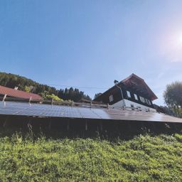 Eine Solarpanelanlage auf einem grasbewachsenen Hügel mit einem Haus im Hintergrund. Das Haus hat ein rotes Dach und mehrere Fenster. Die Solarmodule sind auf einem Holzrahmen montiert.