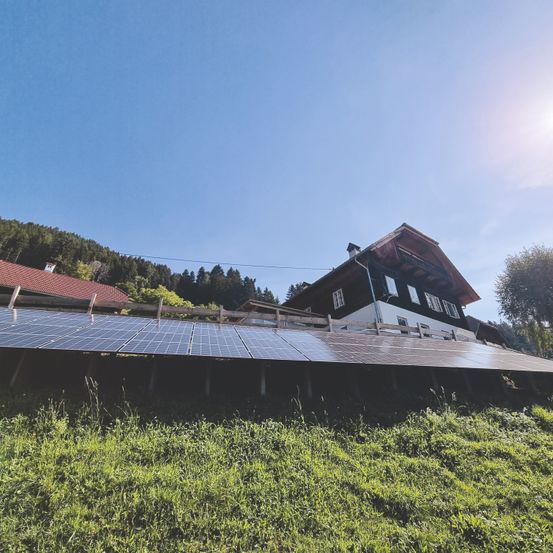 Eine Solarpanelanlage auf einem grasbewachsenen Hügel mit einem Haus im Hintergrund. Das Haus hat ein rotes Dach und mehrere Fenster. Die Solarmodule sind auf einem Holzrahmen montiert.