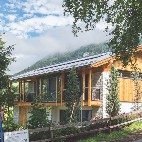 Ein modernes Holzhaus mit Balkon und Solarpanelen auf dem Dach, umgeben von Bäumen und Blumen, in ländlicher Umgebung gelegen.