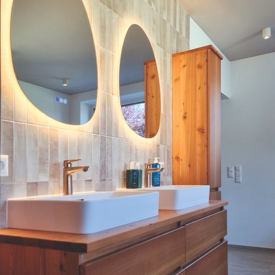 Ein Badezimmer mit zwei Waschbecken, Spiegeln und Holzschränken. Der Waschbeckenbereich ist gut beleuchtet mit dekorativer Beleuchtung.