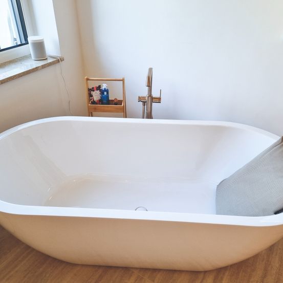 Eine große weiße Badewanne steht auf einem Holzboden. Ein Wasserhahn und ein kleiner hölzerner Regal mit Toilettenartikeln sind in der Nähe. Ein graues Kissen ruht in der Wanne, und ein Fenster mit einem Lautsprecher ist an der Wand zu sehen.