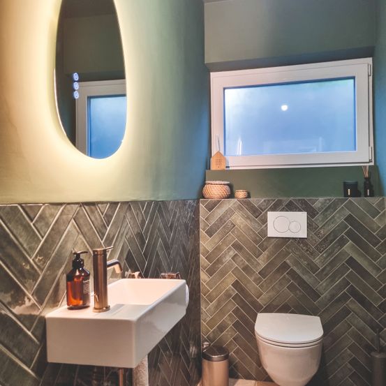 Ein Badezimmer mit einem weißen Waschbecken, einem Spiegel, einer Toilette und einem Fenster. Die Wände sind mit einem Fischgrätmuster verziert.