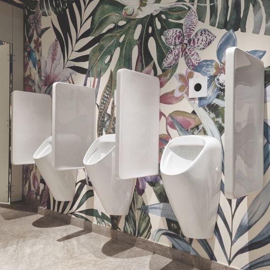Ein modernes Badezimmer mit weißen Urinals an einer Wand mit tropischem Tapetendesign, das Blätter und Blumen zeigt.