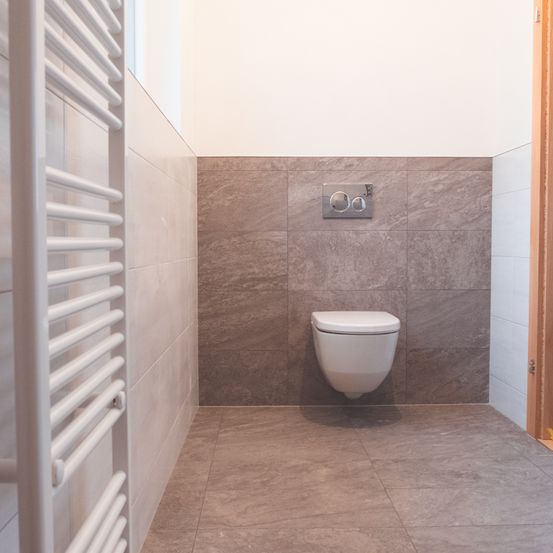 Eine Ecke eines Badezimmers mit einer weißen Toilettenschüssel und grauen Fliesen an den Wänden und am Boden. Ein weißer Handtuchhalter ist an der linken Wand montiert.