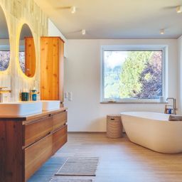 Ein modernes Badezimmer mit Holzböden, einer großen Badewanne, zwei Waschbecken und einem Holzschrank. Es gibt zwei Spiegel und ein Fenster mit Blick auf Bäume.