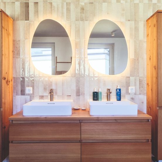Ein Badezimmer mit Holzschränken, zwei weißen Waschbecken und zwei ovalen Spiegeln mit warmer Beleuchtung. Die Wand ist mit einem strukturierten Fliesenmuster verkleidet.