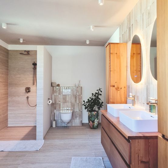 Ein modernes Badezimmer mit Holzwänden, gefliesten Böden und einer Dusche. Eine Toilette und ein Waschbecken mit Wasserhahn sind vorhanden. Ein Holzschrank steht rechts, mit einer Pflanze und Spiegeln darüber.