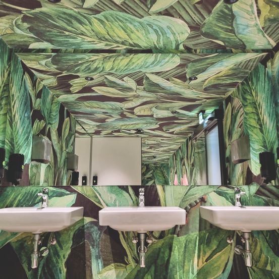 Ein Badezimmer mit tropischem Blatttapeten, drei Waschbecken und einem Spiegel. Die Waschbeckenarmaturen sind silber. Über den Waschbecken befinden sich Seifenspender.