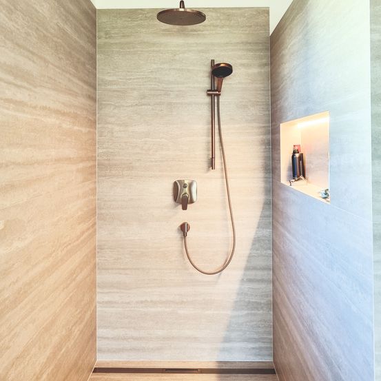 Ein Duschbereich mit einem runden, kupferfarbenen Duschkopf, der an einer Wand befestigt ist. Die Wand ist gefliest und es gibt ein kleines Regal mit Flaschen darin.