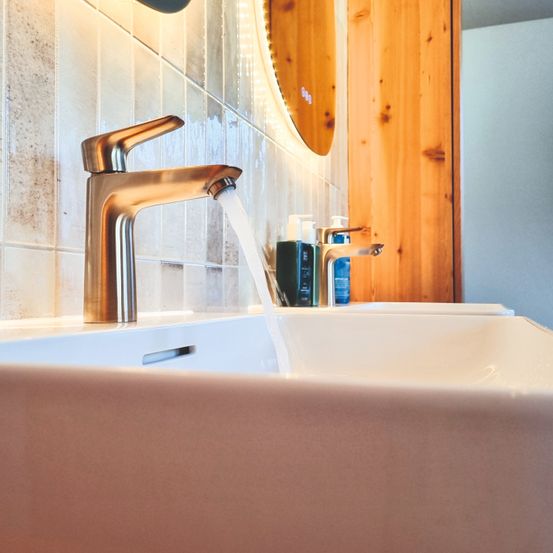 Ein Badezimmer mit einem Waschbecken, einem Wasserhahn, fließendem Wasser und Seifenflaschen auf dem Waschtisch. Die Wand ist gefliest und ein Spiegel ist an der Wand befestigt.