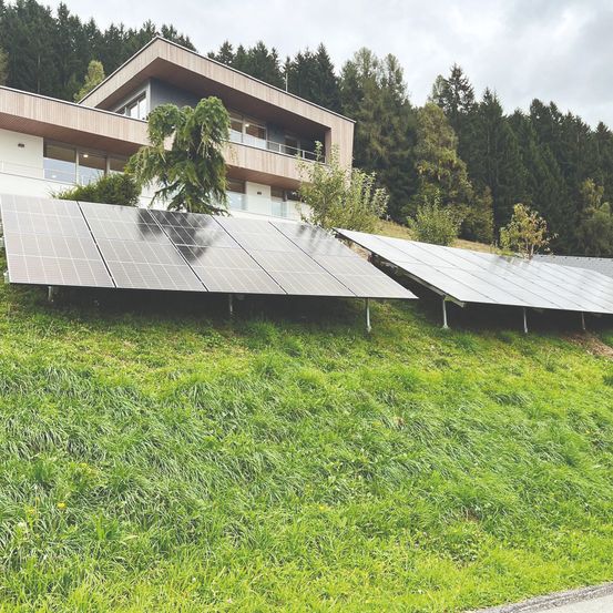 Solarmodule auf einem Hang mit einem modernen Haus und Bäumen im Hintergrund. Das Haus hat einen Balkon und viele Fenster.