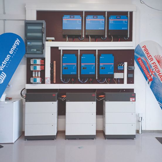 Eine Wand mit drei großen grauen und weißen Schränken, jeweils mit mehreren Tasten und Bildschirmen, ist im Raum zu sehen. Ein blauer Banner mit Text hängt links.