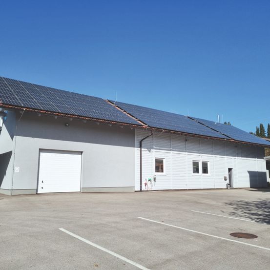 Ein weißes Gebäude mit Solarpanelen auf dem Dach, einer Garagentür und Fenstern. Es ist von einem Parkplatz umgeben.