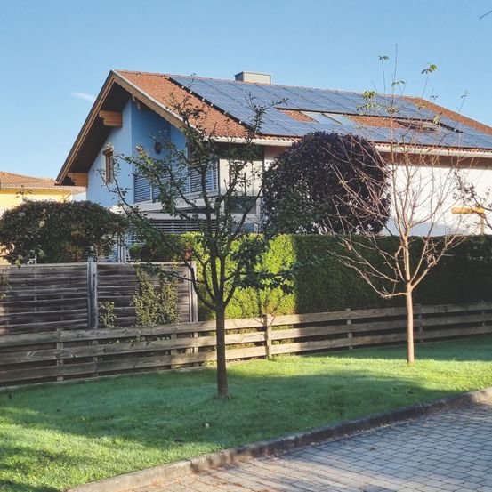 Ein blaues Haus mit einem braunen Dach und Solarmodulen. Im Garten gibt es einen Holzzaun und grünen Rasen. Zwei Bäume sind im Garten gepflanzt.
