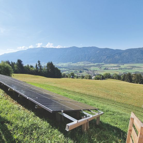 Eine Solarpanel-Anlage ist auf einem Grashügel mit Blick auf ein Tal und Berge unter einem klaren Himmel positioniert.