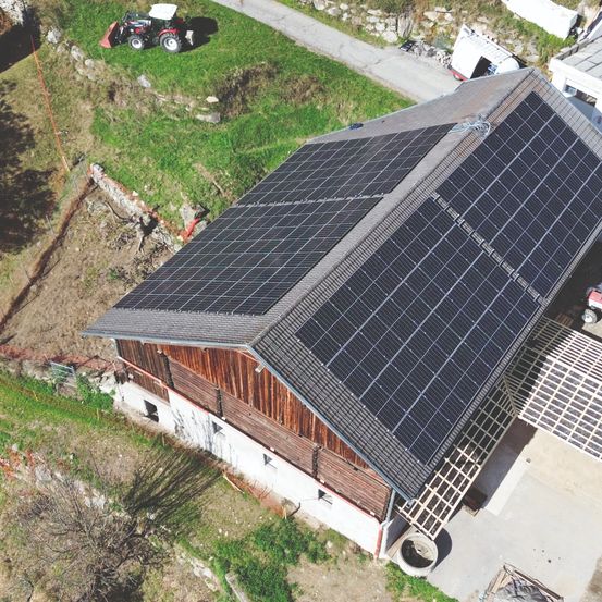 Luftaufnahme eines Gebäudes mit einer großen Solarpanelanlage auf dem Dach. In der Nähe ist ein Traktor auf einem Grasfeld geparkt.
