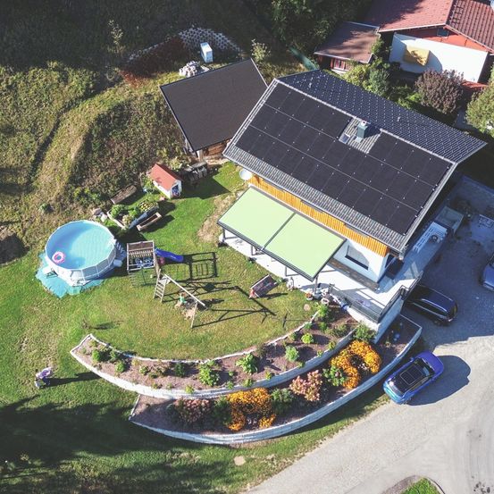 Luftaufnahme eines modernen Hauses mit Solarmodulen, einem Garten, einem Pool und einem Spielplatz in einem grünen Bereich.