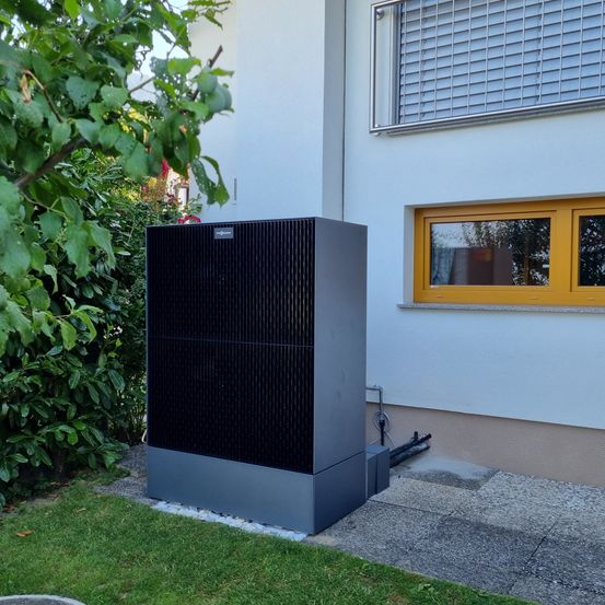 Ein großes schwarzes und graues Außengerät steht auf dem Betonpflaster neben einem weißen Haus mit gelben Fenstern.